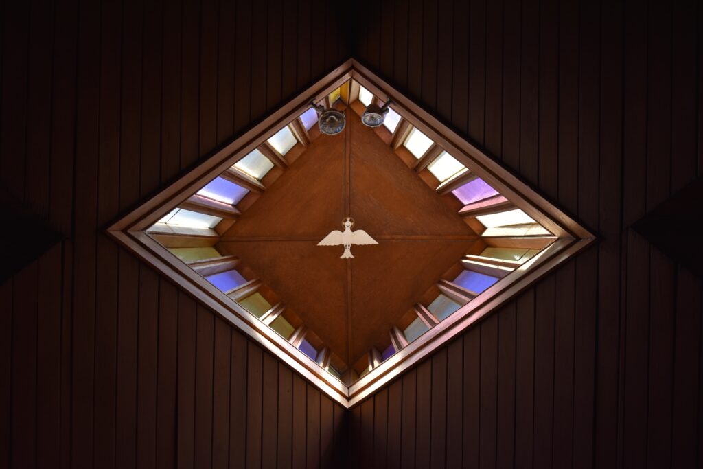 An einer Kirchendecke hat es eine Taube. Ringsherum sind bunte Glasfenster.