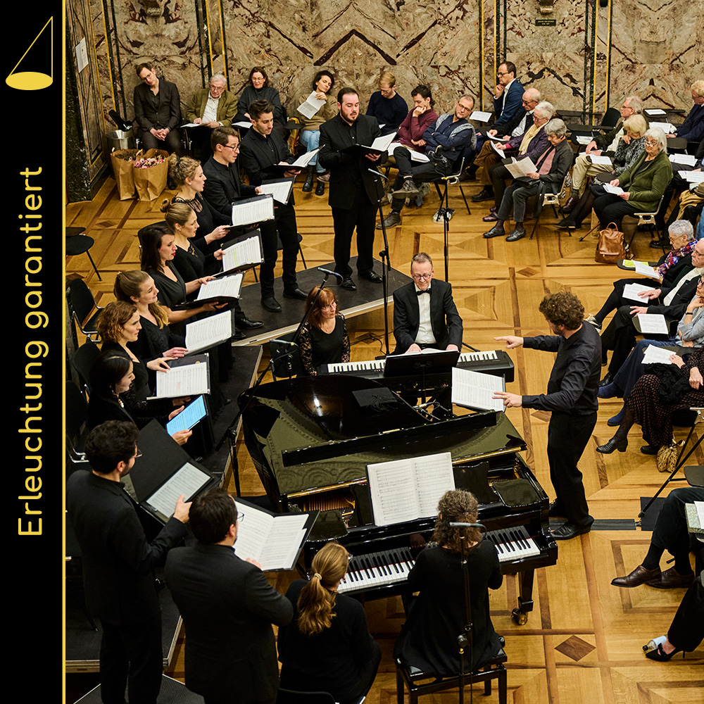 Konzertaufführung von schräg oben fotografiert. Im Zentrum ein Flügel, ein Klavier und der Dirigent, links im Bild ein Chor (eine Reihe) aus 12 Personen, rechts und im Hintergrund ist ein Teil des Publikums zu sehen