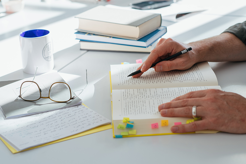 Zwei Hände ruhen auf einem Fremdsprachenwörterbuch, daneben steht eine Kaffeetasse, liegen weitere Bücher und eine Brille