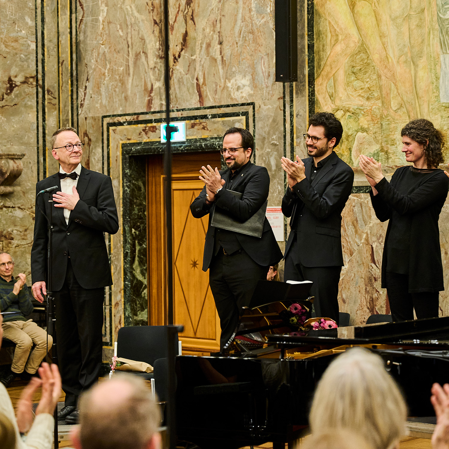 Dr. h.c. Rudolf Lutz (ganz links) bedankt sich nach dem Konzert mit einer Geste (seine linke Hand liegt auf seiner Brust), rechts sind drei Sänger:innen zu sehen, die ihm applaudieren (im Vordergrund unscharf ein paar Köpfe aus dem Publikum)